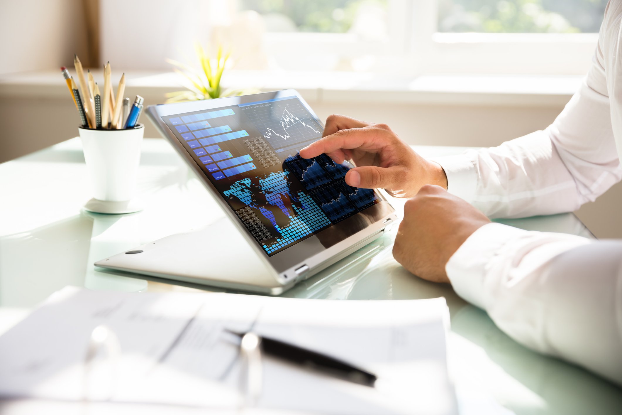 A businessman's hands touching a financial chart on a laptop, which is sitting on a desk with a pencil holder nearby.