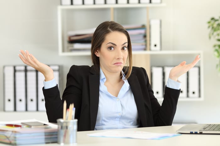 Young woman in business attire, sitting at a desk with a confused demeanor.