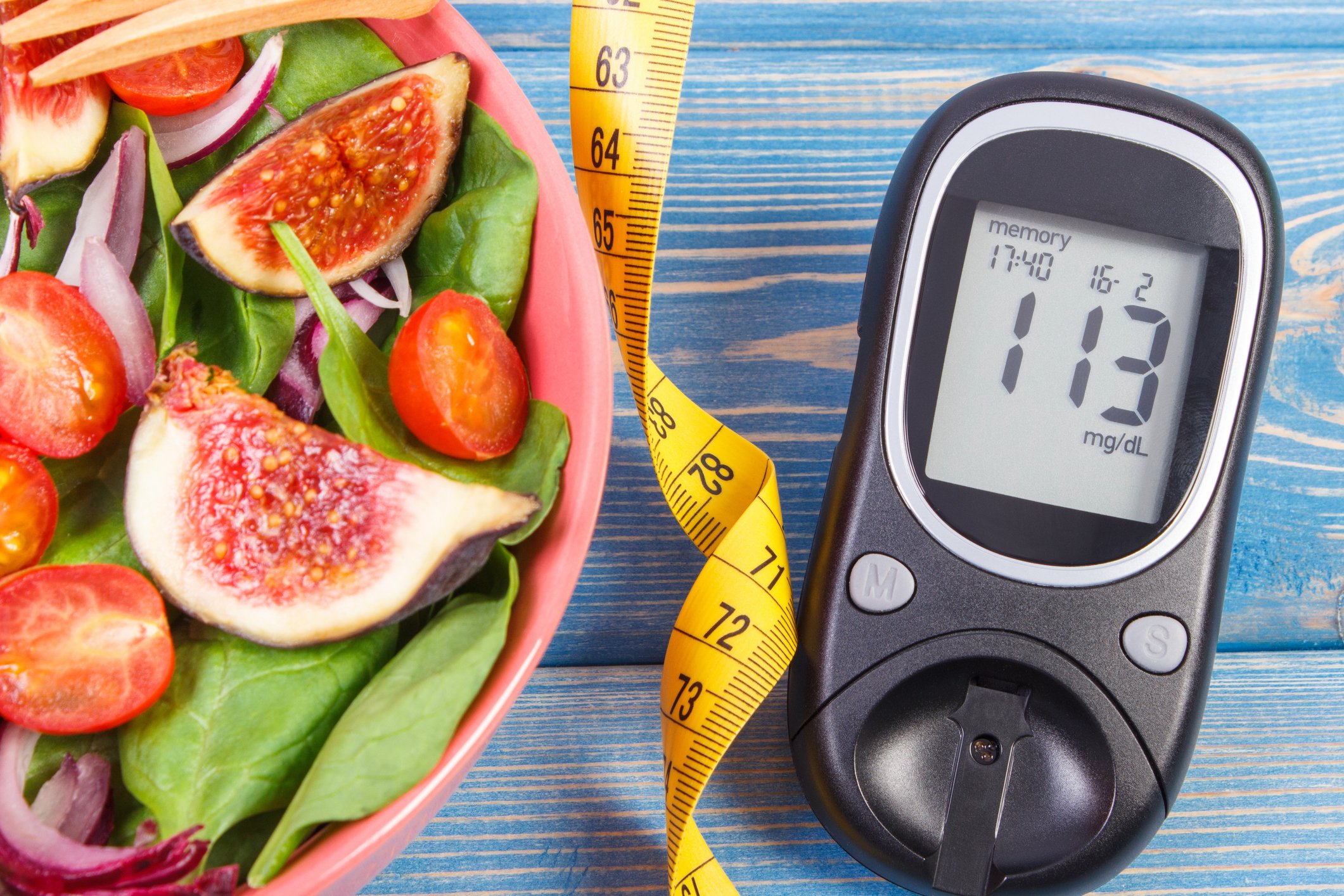A blood glucose monitor sits next to a measuring tape and a bowl of salad