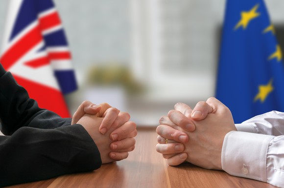 Two people sitting at a table facing each other, with a U.K. flag and a European Union flag behind them