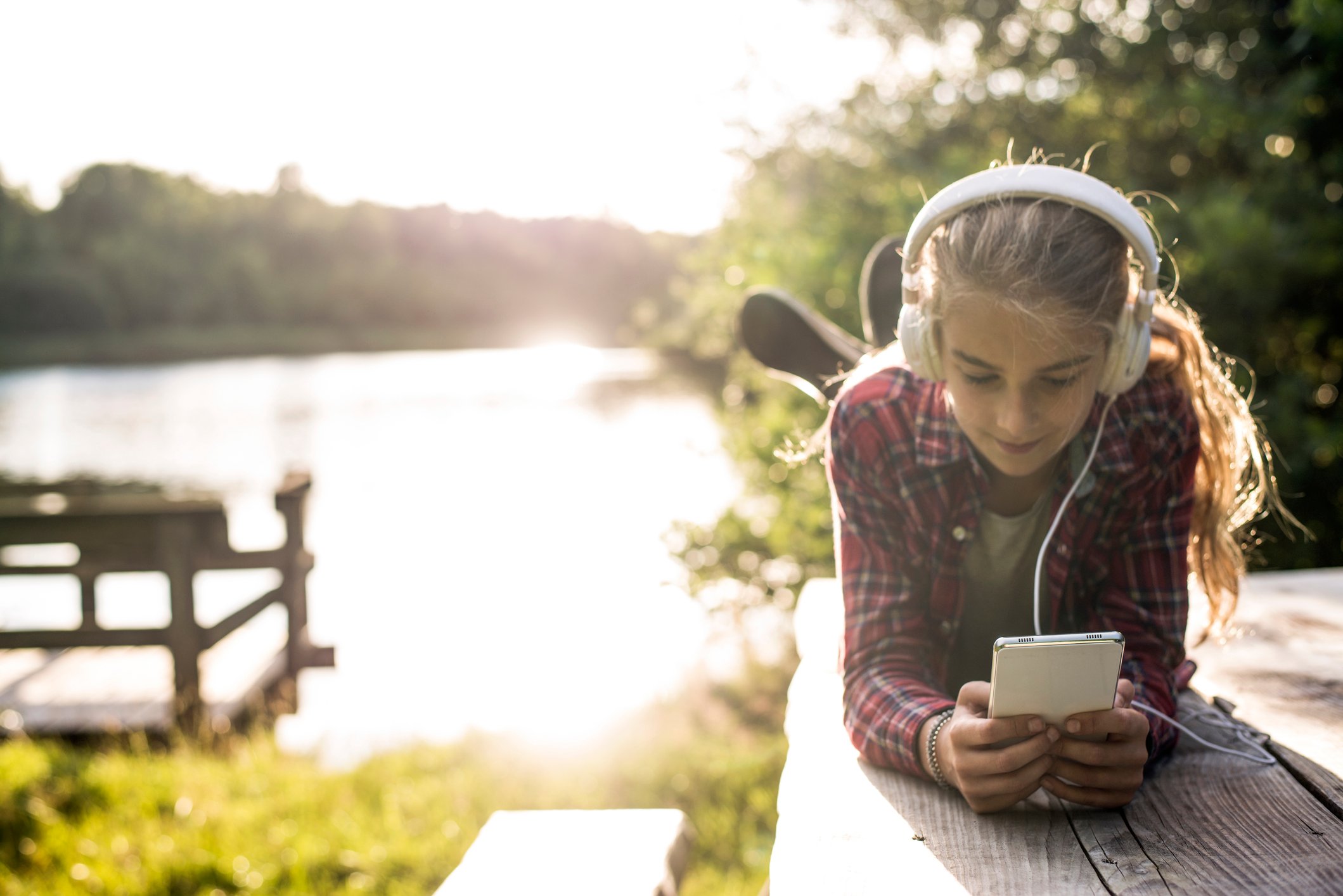 A girl laying on a picnic table near a lake, wearing headphones and using a smartphone.