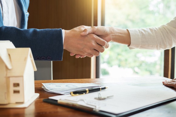 Two people shaking hands over a clipboard on top of a desk.