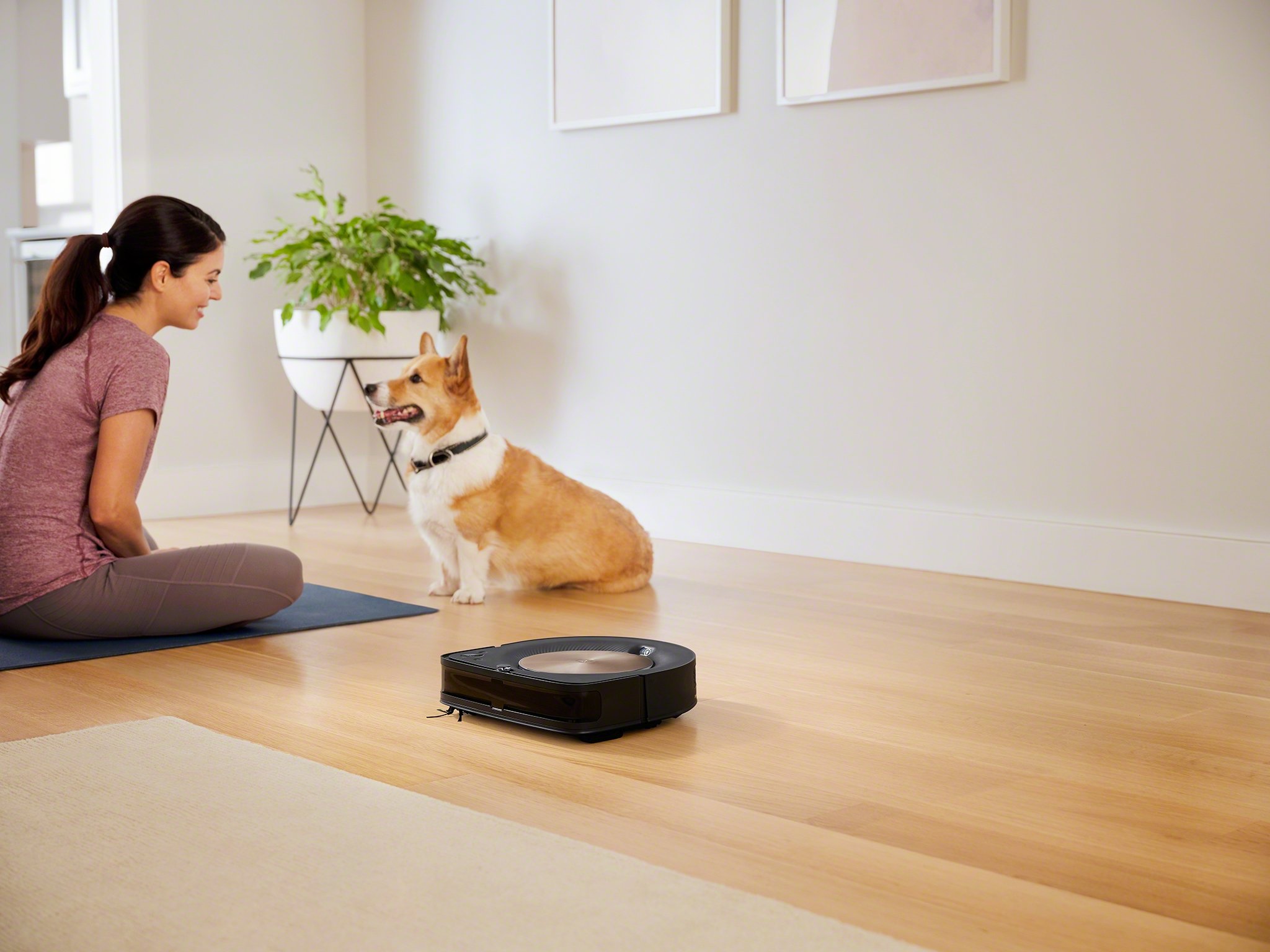 A woman sitting on the floor looking a small dog as a a robotic vacuum cleans in the foreground