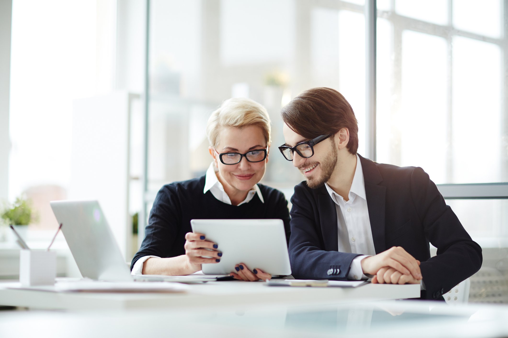 A man and woman in an office setting look smiling at the screen of a tablet computer.