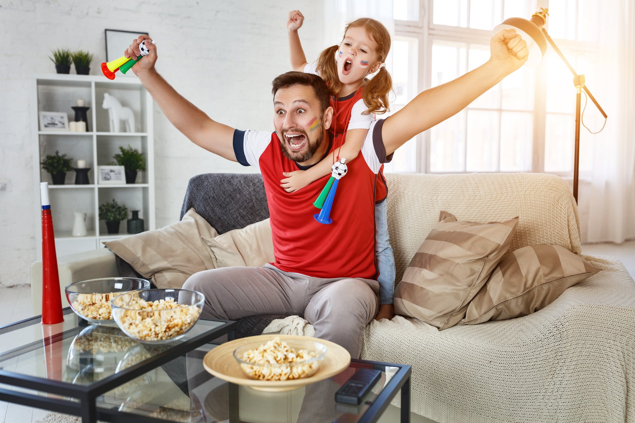 A father and his young daughter, with painted faces, cheering on their favorite team from their couch, with popcorn on a table in front of them. 
