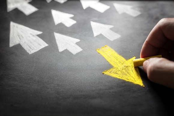 A hand drawing a yellow arrow in chalk that is opposing the direction of many other white arrows.