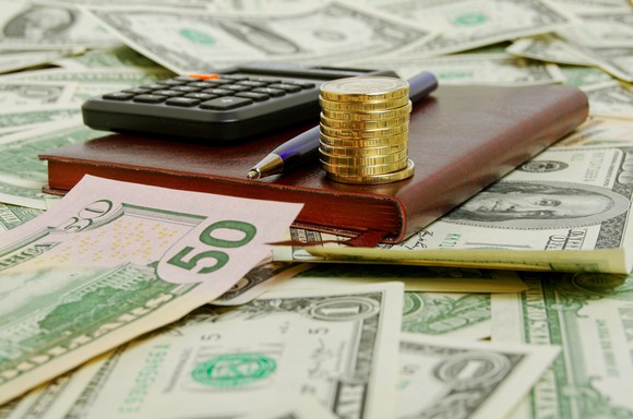 A calculator, a pen, and a stack of coins sit atop a scattered pile of U.S. currency.