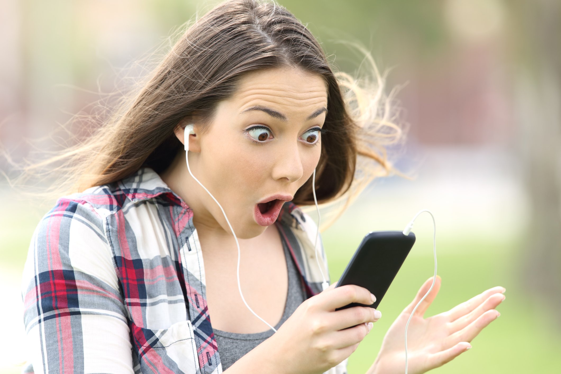 A young woman wearing headphones stares in wide-eyed amazement at her phone.