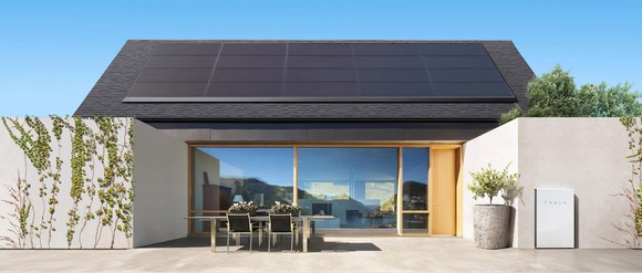 A roof with a solar panel installation on it with a Tesla-branded storage unit on the ground on the lower-right. 