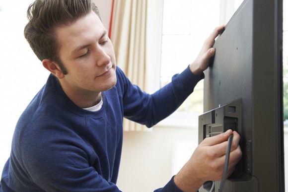 A young man plugs a coaxial cable into the back of a television.