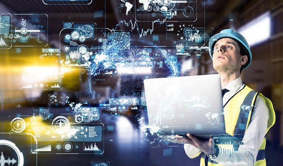 Man wearing hard hat and fluorescent yellow vest holding laptop and looking at various projected Internet of Things icons