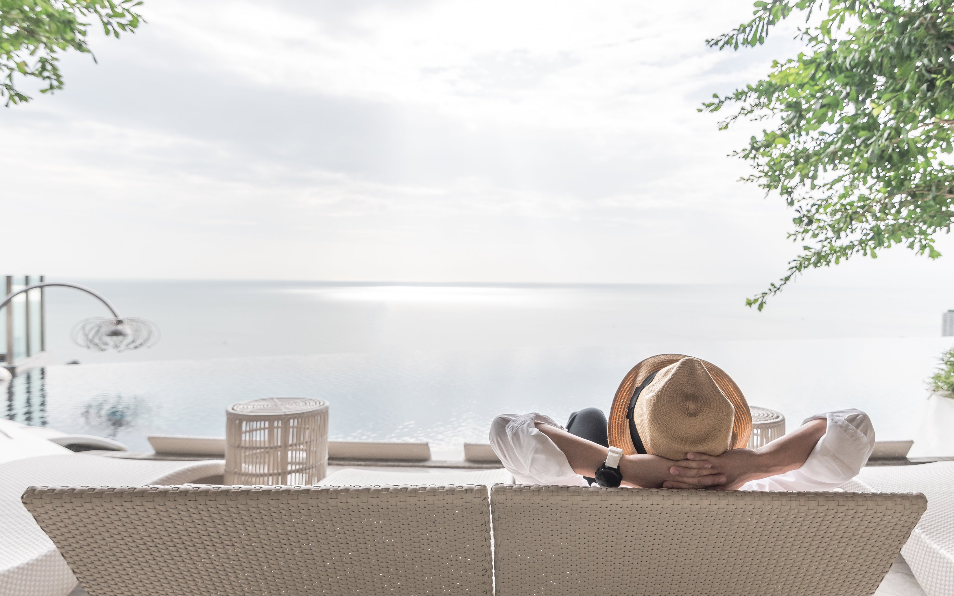 Seen from behind, a woman relaxes on a wicker chair facing the water at an upscale beach resort.