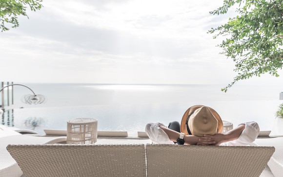 Seen from behind, a woman relaxes on a wicker chair facing the water at an upscale beach resort.