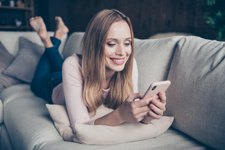 Young woman lounging on a tan sofa while using a smart phone.