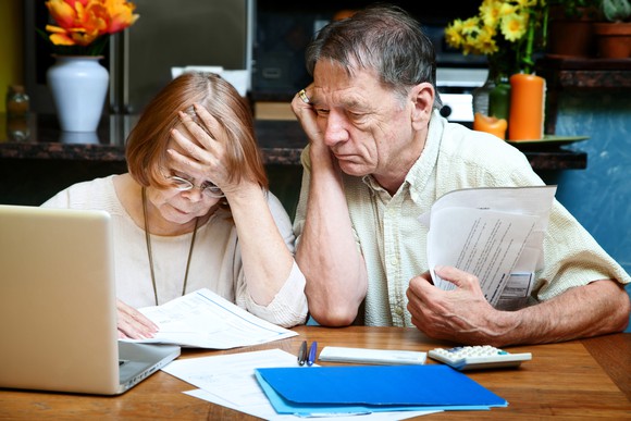 Older man and woman sitting at table with laptop and documents in front of them, looking concerned