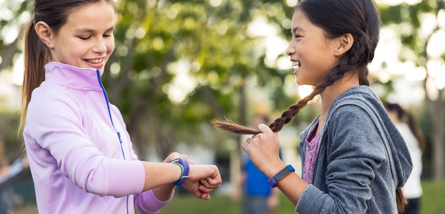 Two smiling girls with wearable devices on their wrists, in an outdoor setting.
