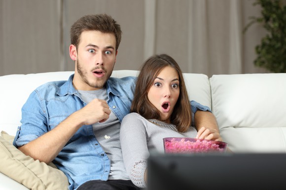 A young couple sharing a bucket of popcorn on their living room couch, staring at the TV screen in wide-eyed amazement.