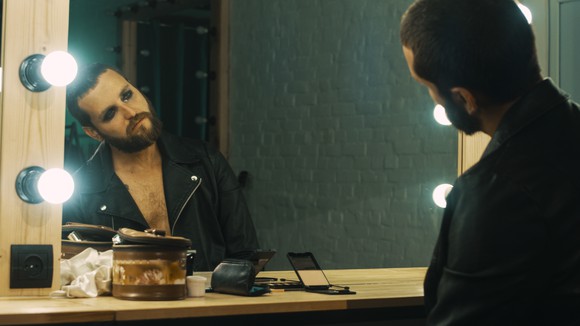 A rock star looking in a mirror, head tilted sideways with a tired look on his makeup-heavy face.