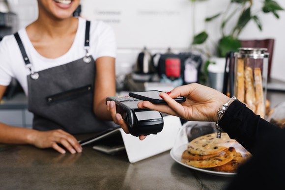 A woman's hand holding a smartphone and making a mobile payment to a server behind the counter at a coffee shop.