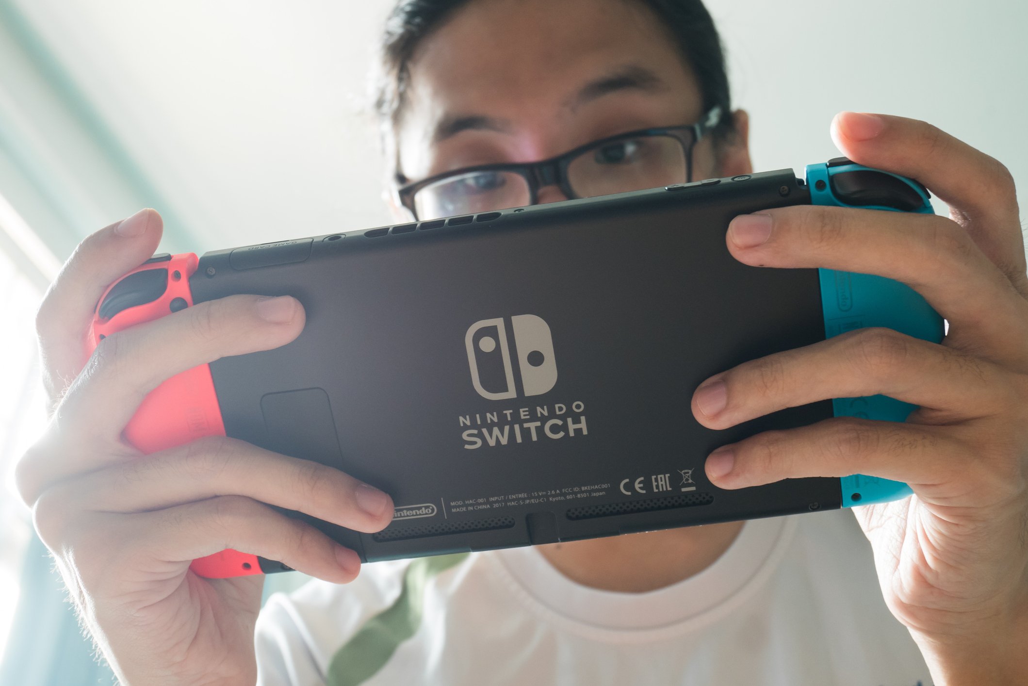 A young gamer in glasses holds his Nintendo switch viewed from below with his eyes peering over. 