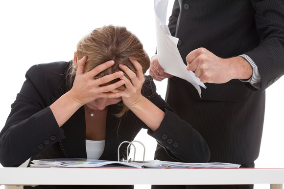 Seated woman covering her head while man in suit stands over her while holding a stack of papers.