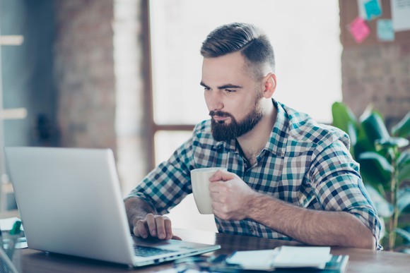 A man holds a cup of coffee while using his laptop.