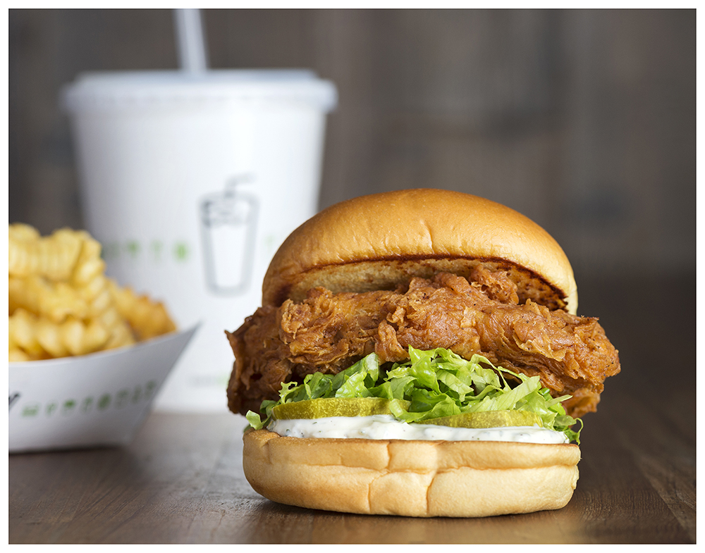 A fried chicken sandwich from Shake Shack. A drink and box of fries are displayed in the background.