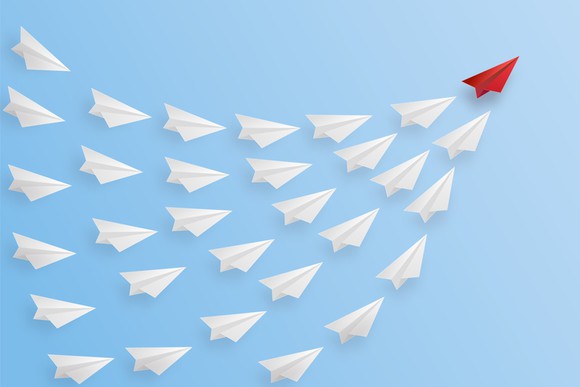 A red paper airplane leading a rising swarm of white paper airplanes.