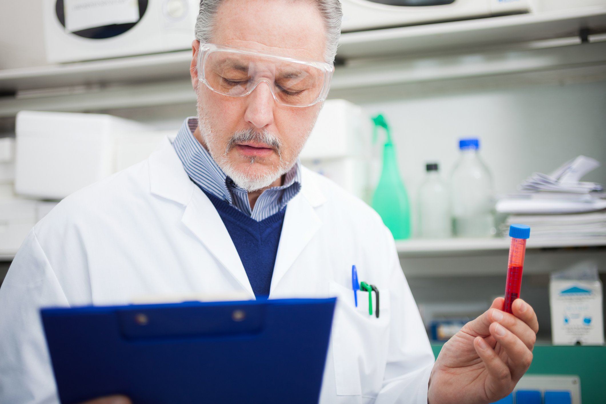 A biotech lab technician holding a blood sample in his left hand while reading from a blue clipboard in his right hand.