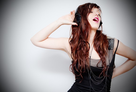 A smiling young woman with headphones dances with one hand cupped to her ear.