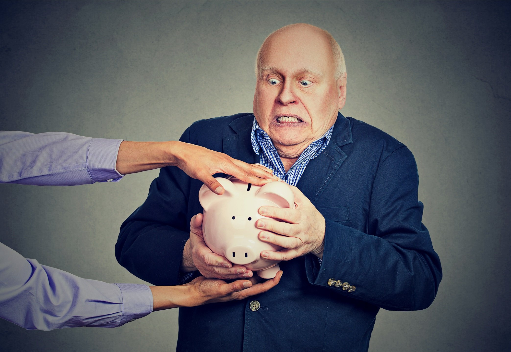 Older man with frightened expression holding on to his piggy bank, while a pair of hands tries to take it away.