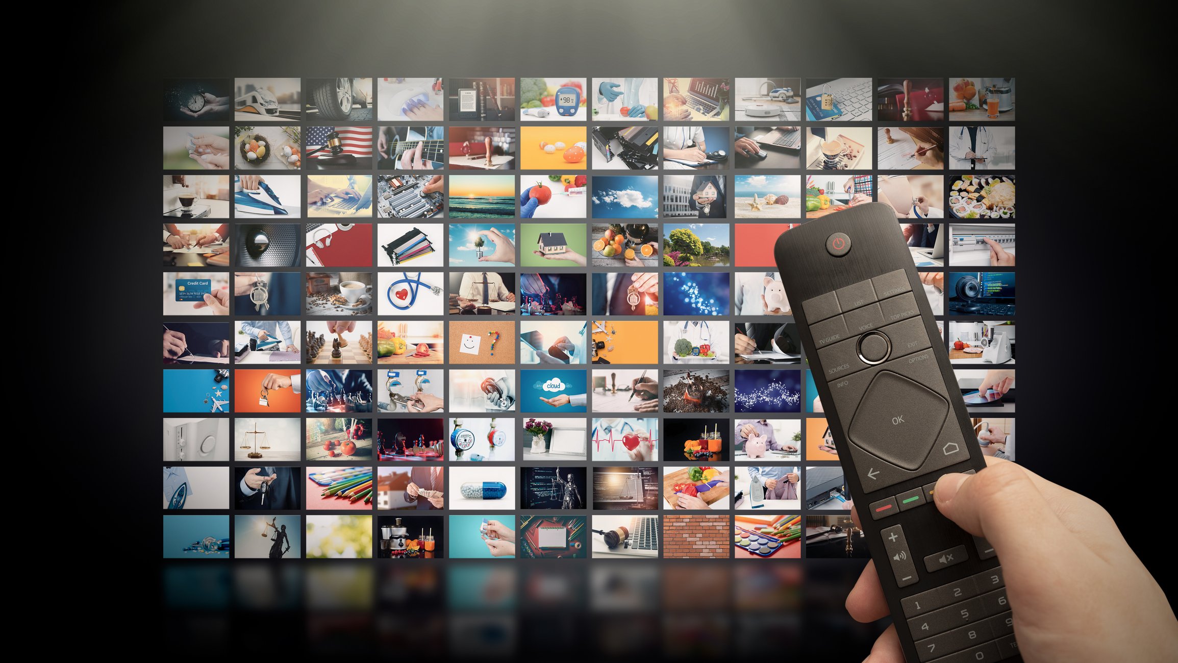 Close-up shot of a hand holding a TV remote in front of a wall made up of 120 TV screens, all showing different content.