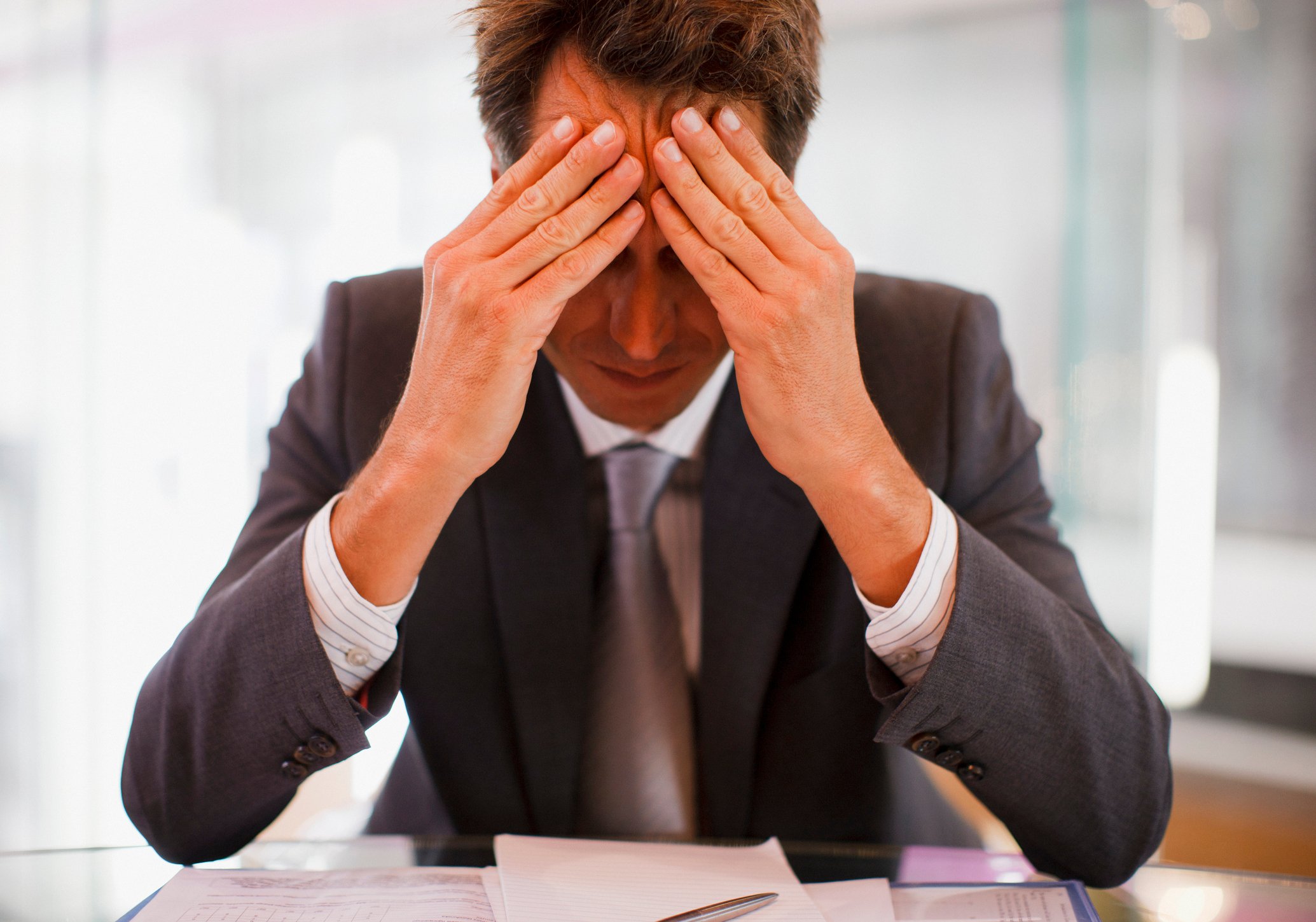 Man in suit holding his head while seated in front of documents