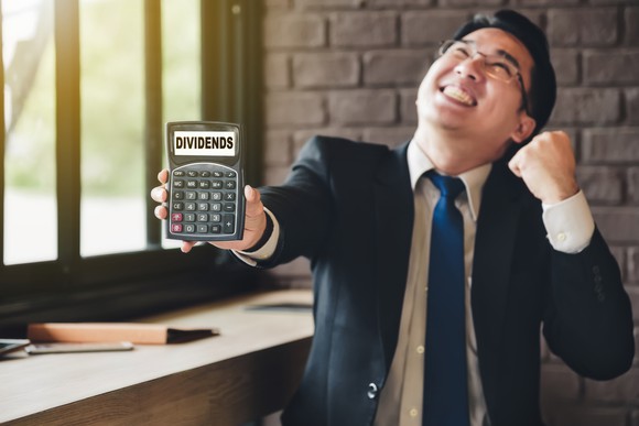 A young man in a business suit pumps his fist smiles and holds out a calculator that says the word dividends on its screen. 