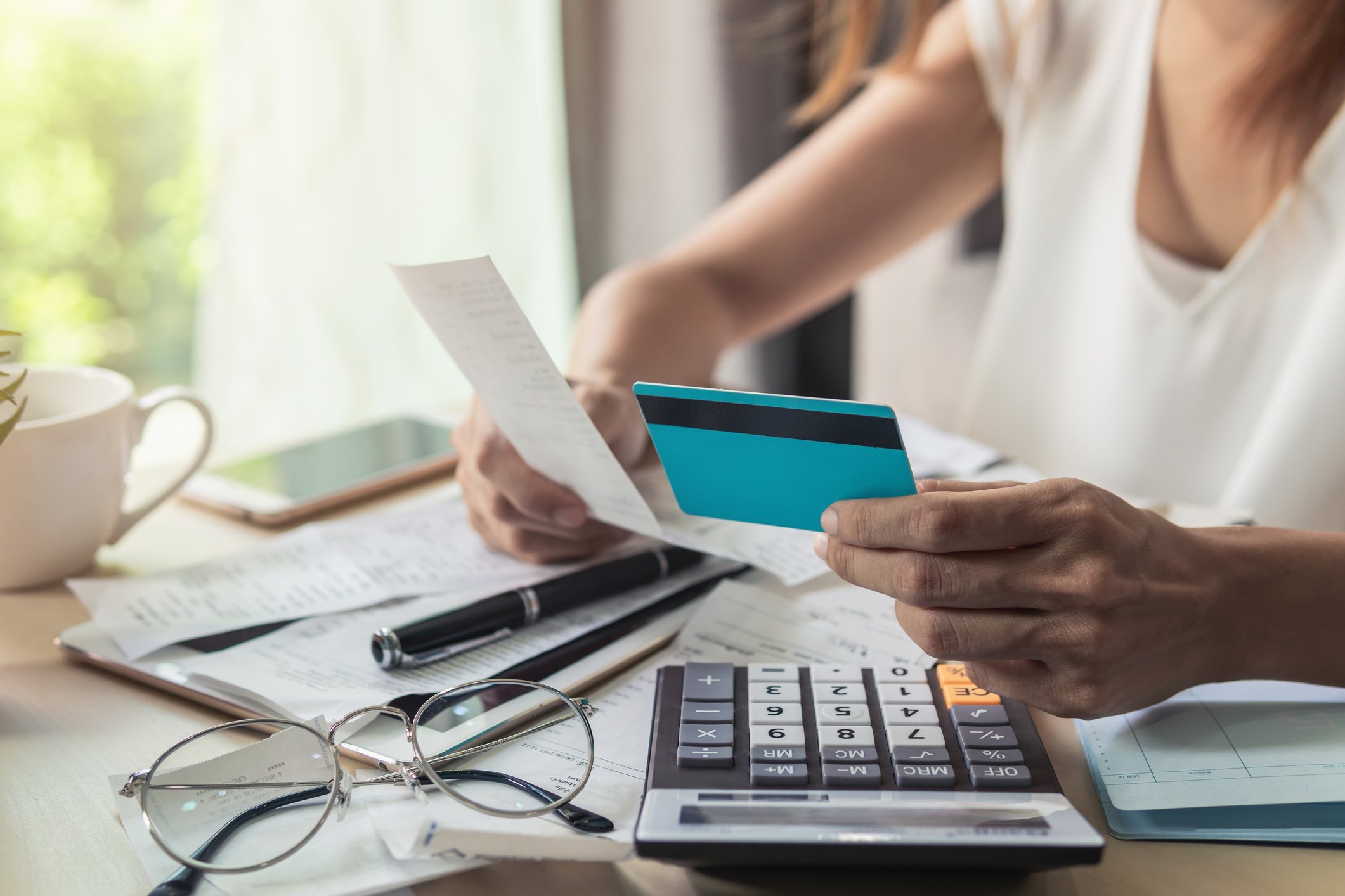 Person sitting at a desk holding a credit card and receipt, with a calculator and papers strewn on the desk.