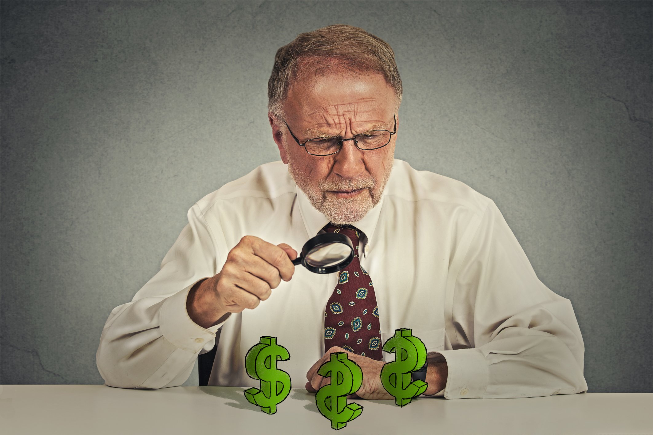 A senior man using a magnifying glass to closely examine dollar signs on the table in front of him.