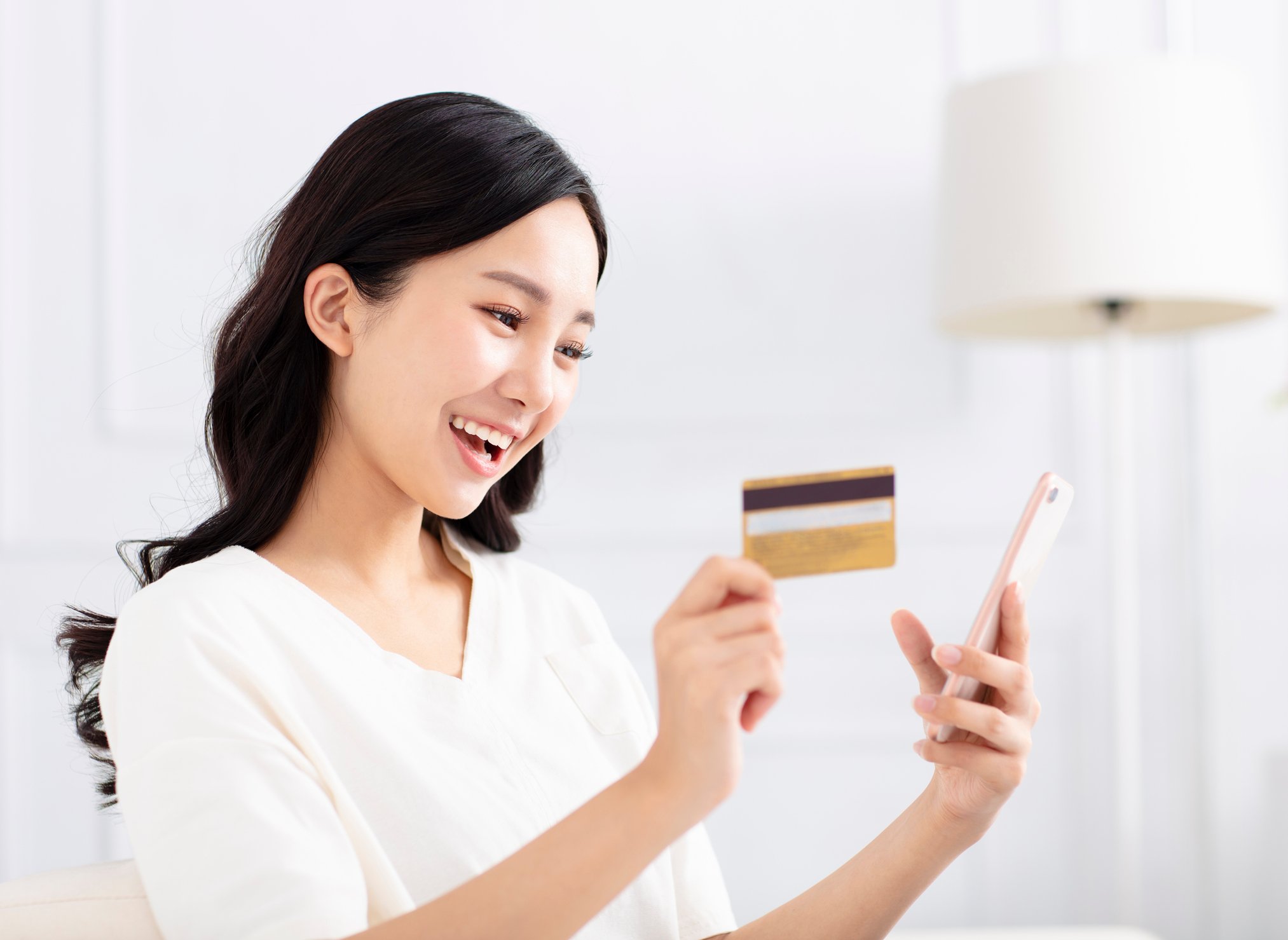 A smiling young Chinese woman with her smartphone in one hand and a credit card in the other.