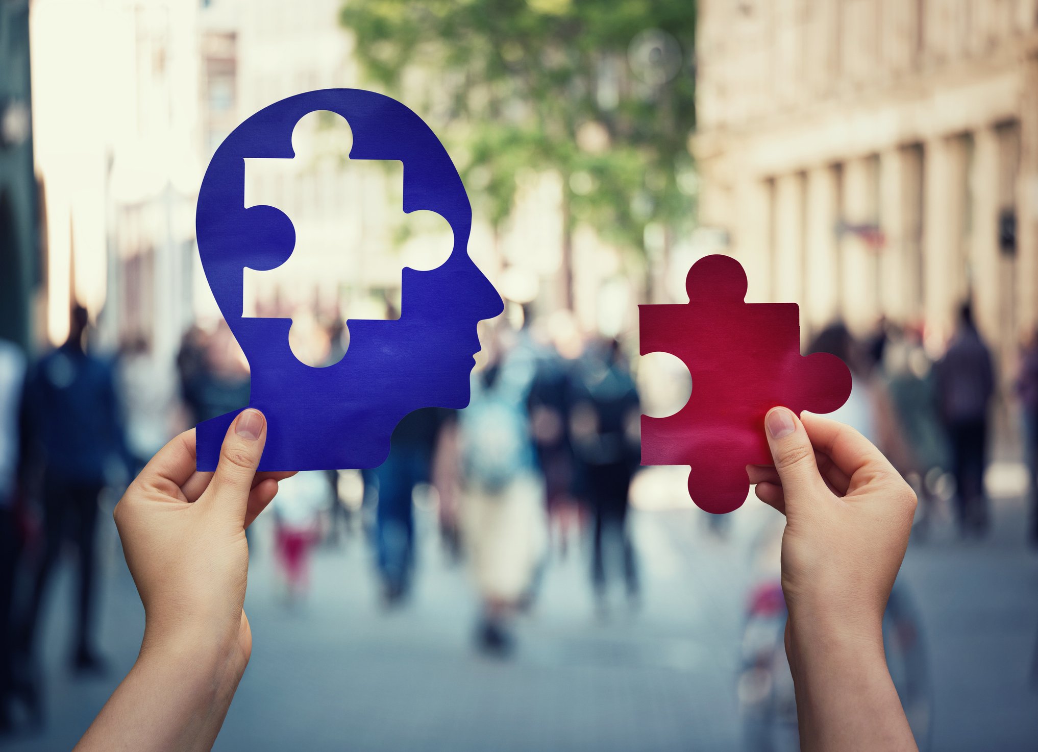 Hand holding a blue figure of a head with a jigsaw puzzle piece cut out of it and the person's other hand holding a red jigsaw puzzle piece that fits into the cutout space in the blue head, with people walking on a city street in the background.