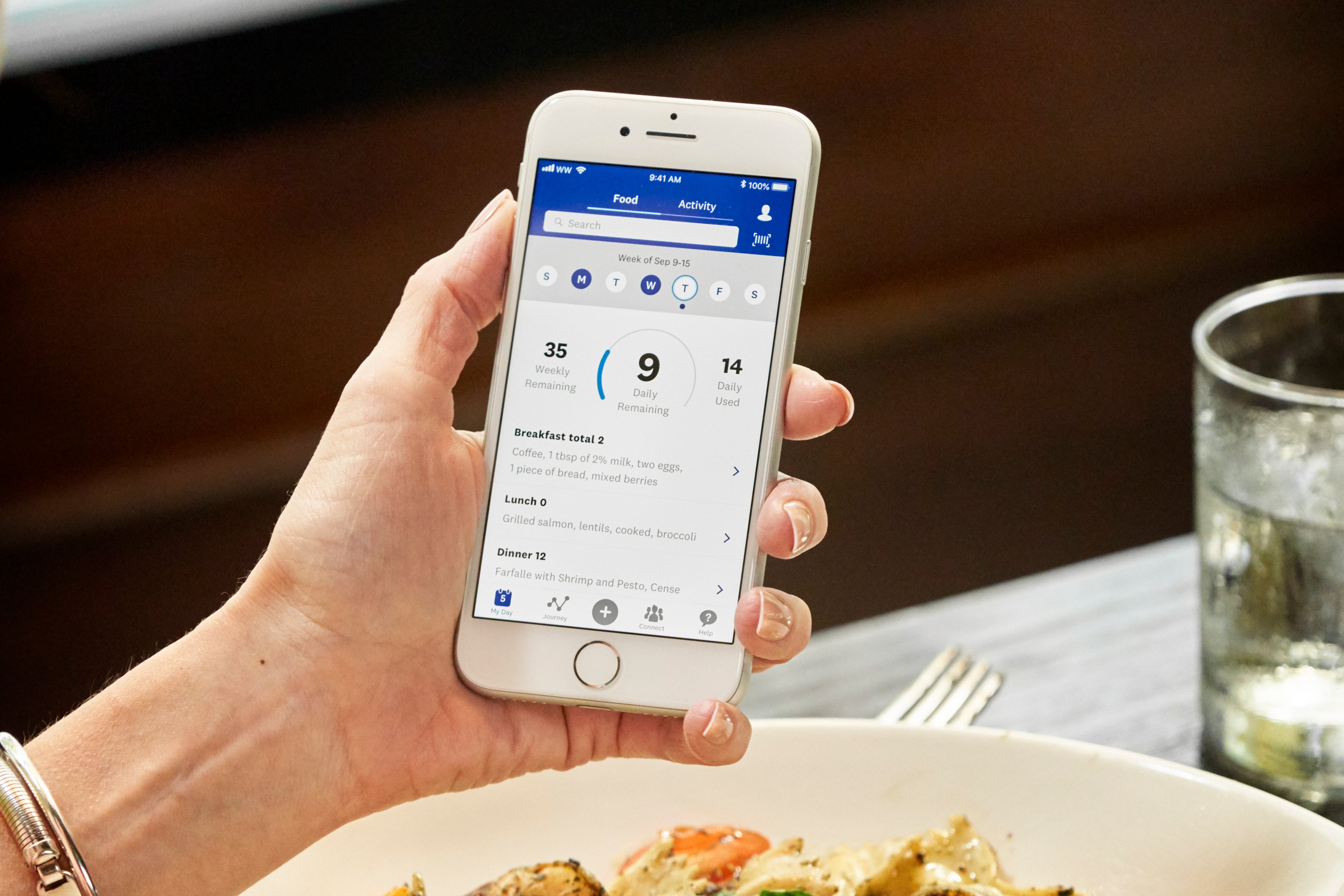Hand holding phone (over plate of food and drink) with WW app displayed on screen.