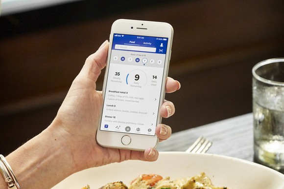 Hand holding phone (over plate of food and drink) with WW app displayed on screen.