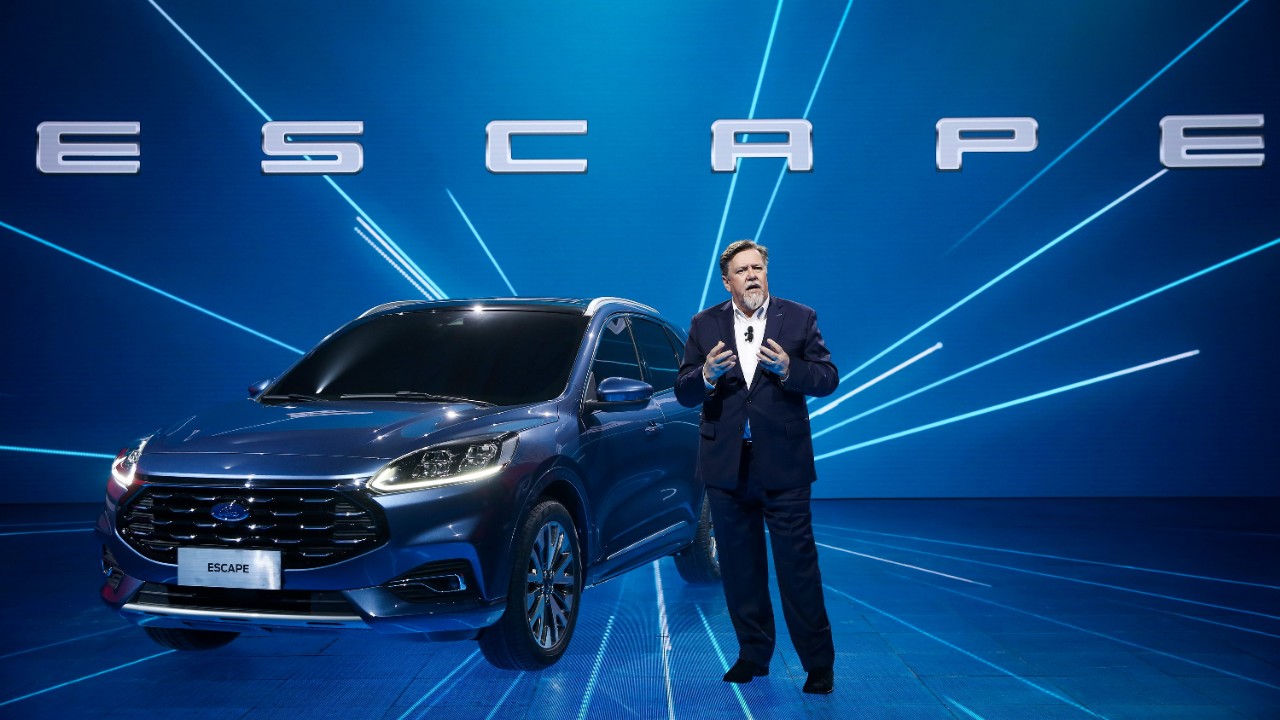 A man standing next to a blue Ford Escape SUV on a blue stage. 
