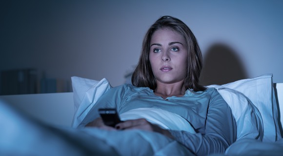 A young woman lies in bed watching TV and holding her phone. 
