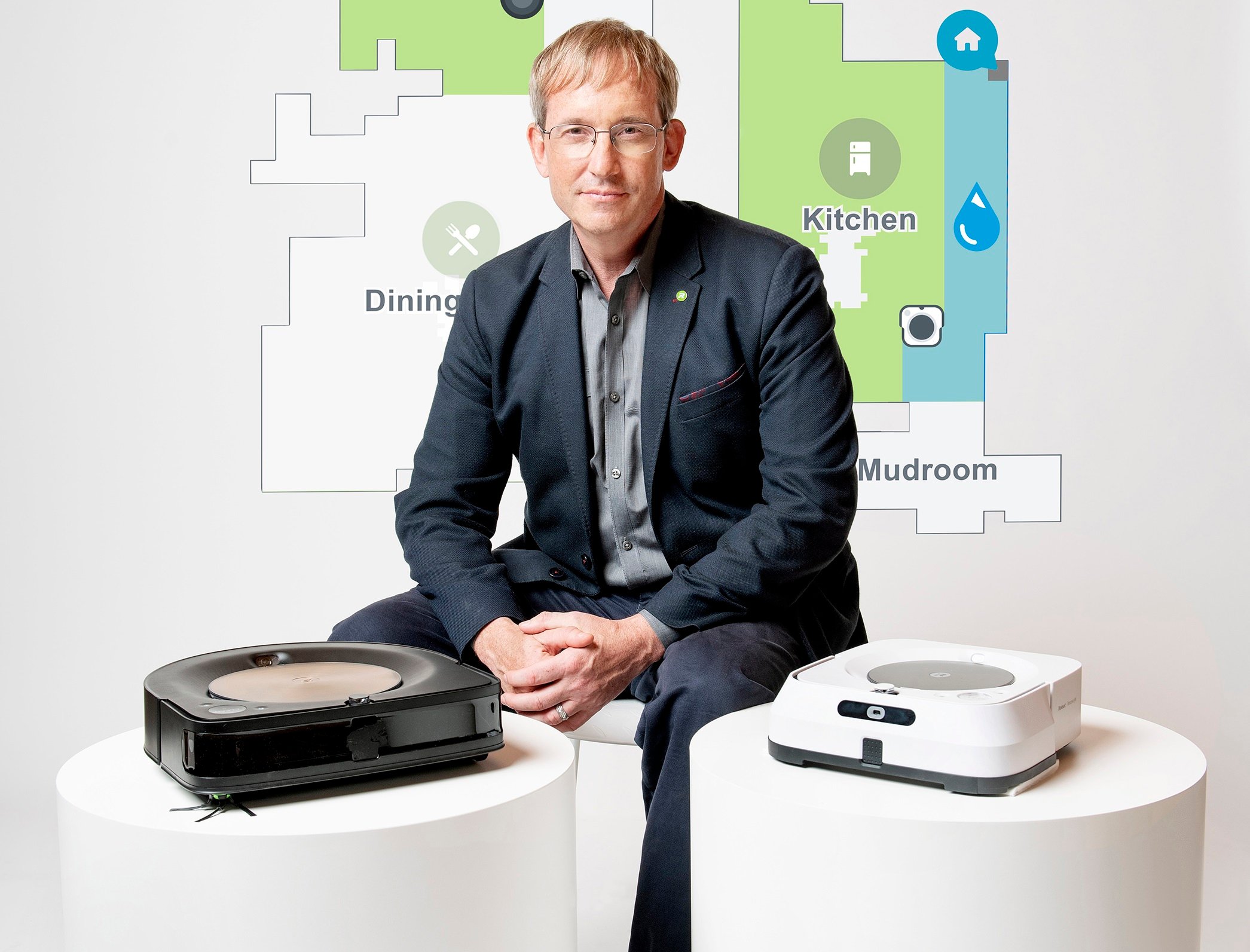 iRobot chairman, co-founder, and CEO Colin Angle sitting next to high-end Roomba and Braava robots with a house map in the background.