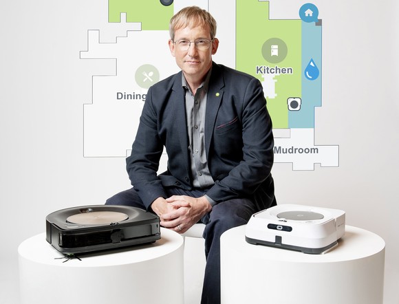 iRobot chairman, co-founder, and CEO Colin Angle sitting next to high-end Roomba and Braava robots with a house map in the background.