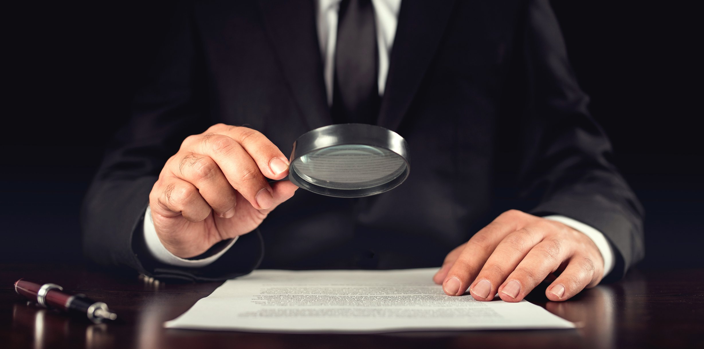 A businessman in suit and tie holds a magnifying glass up to a sheet of paper laying on a wooden desk. 