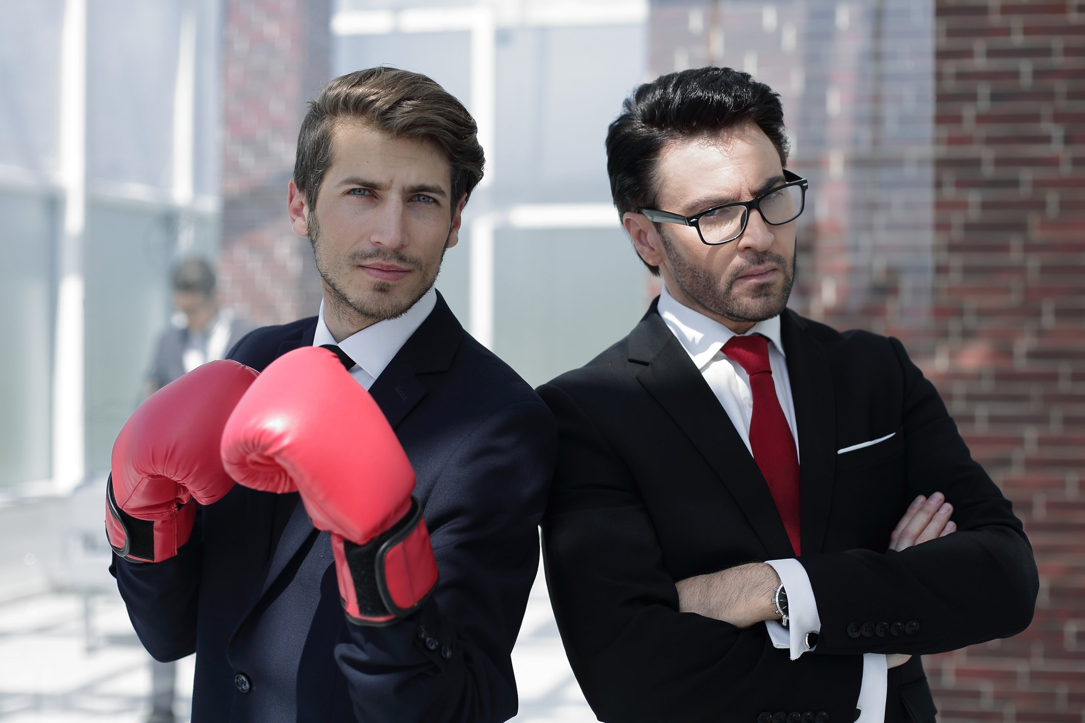 Two young businessmen stand back to back in an alley, one wearing red boxing gloves.