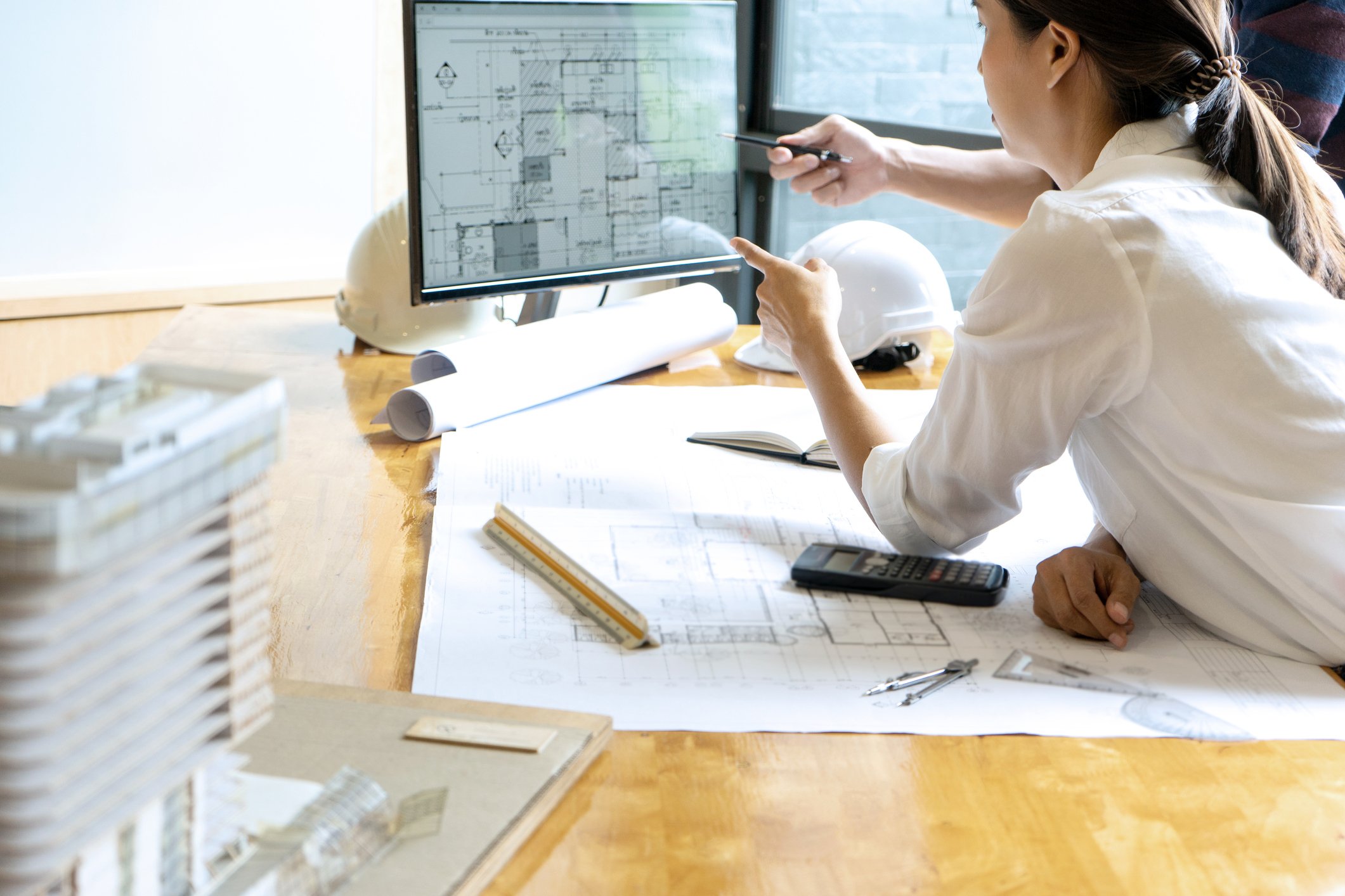 A woman leans on blueprints and points to an architectural rendering on a computer screen.