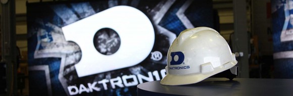 A Daktronics-branded white helmet sits on a table in front of a large display showing a much bigger version of the same logo.