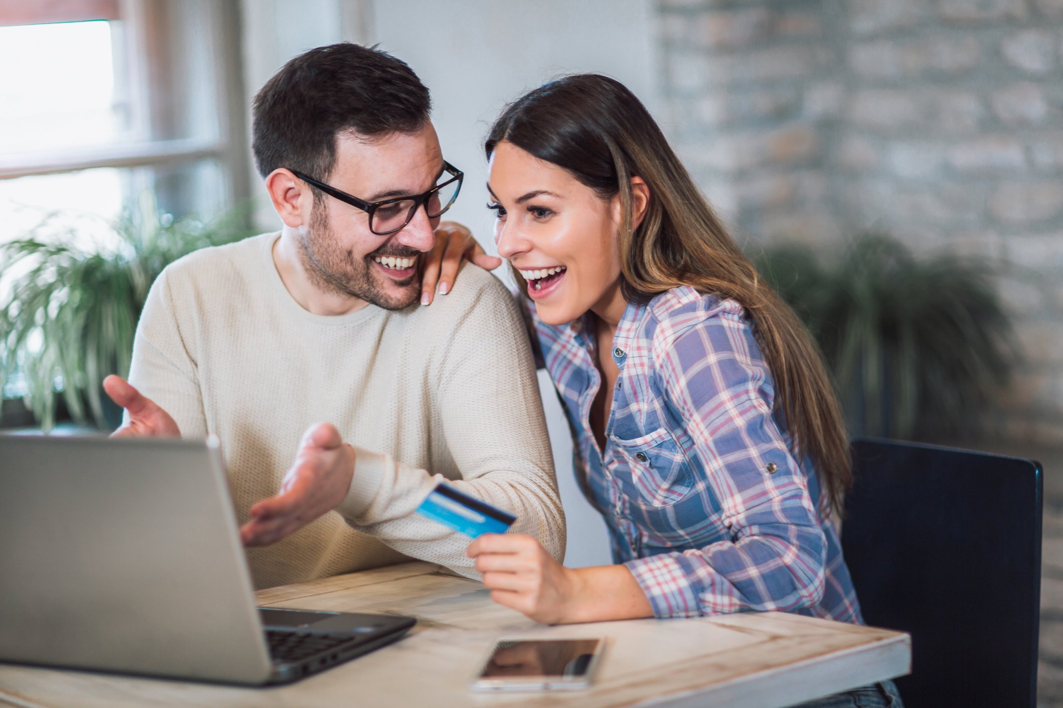 A woman looking at a laptop screen and smiling. She is holding a credit card. A man is gesturing to the laptop screen and smiling.