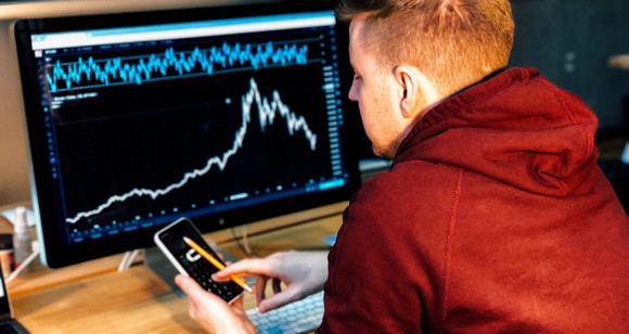 A man scrolling through a smartphone as he sits at a desk with a computer screen displaying a stock chart.